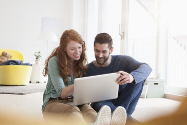 Smiling young couple sitting on floor in the living room using laptop - RBF002836