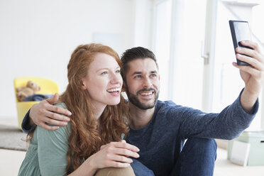 Lächelndes junges Paar macht ein Selfie mit Smartphone im Wohnzimmer - RBF002838