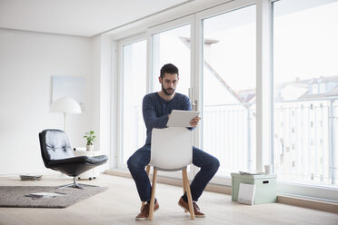 Young man sitting on chair in his living room using digital tablet - RBF002844