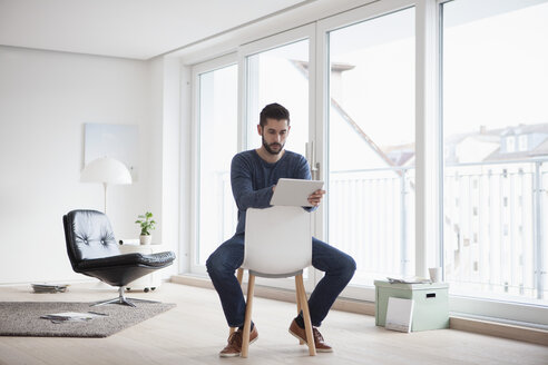 Junger Mann sitzt auf einem Stuhl in seinem Wohnzimmer und benutzt ein digitales Tablet - RBF002844