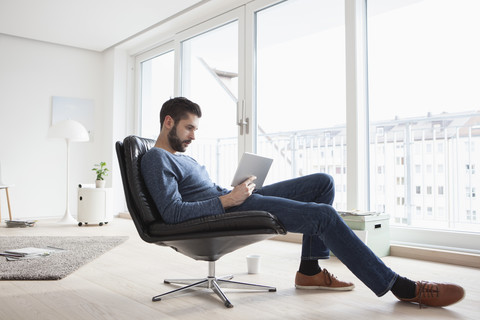 Junger Mann sitzt auf einem Ledersessel in seinem Wohnzimmer und benutzt ein digitales Tablet, lizenzfreies Stockfoto