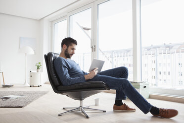 Junger Mann sitzt auf einem Ledersessel in seinem Wohnzimmer und benutzt ein digitales Tablet - RBF002848