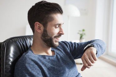 Smiling young man sitting on leather chair looking at his smartwatch - RBF002766