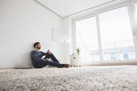 Junger Mann sitzt auf dem Boden des Wohnzimmers und schaut durch das Fenster, lizenzfreies Stockfoto