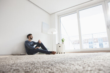 Junger Mann sitzt auf dem Boden des Wohnzimmers und schaut durch das Fenster - RBF002849