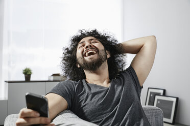 Laughing young man with smartphone sitting on couch at home - PDF001074
