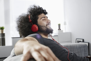 Junger Mann sitzt zu Hause auf der Couch und hört mit Kopfhörern Musik - PDF001075