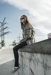 Young woman wearing sunglasses sitting on a wall with smartphone - UUF004587