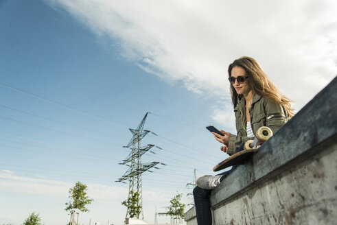 Junge Frau mit Skateboard und Smartphone hört Musik mit Kopfhörern - UUF004588