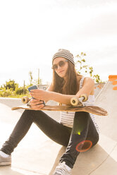 Porträt eines jungen Skateboarders mit Sonnenbrille, der in einem Skatepark sitzt und sein Smartphone benutzt - UUF004600