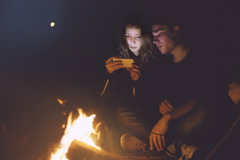 Junges Paar sitzt zusammen am Lagerfeuer und schaut auf sein Handy, lizenzfreies Stockfoto