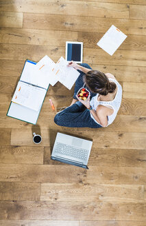 Student sitting on wooden floor surrounded by papers, laptop, digital tablet, file folder and coffee - UUF004699