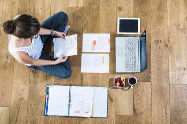 Student sitting on wooden floor surrounded by papers, laptop, digital tablet, file folder, coffee and fruit bowl - UUF004700