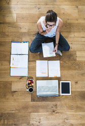 Student sitting on wooden floor surrounded by papers, laptop, digital tablet, file folder, coffee and fruit bowl - UUF004701