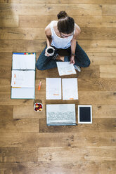 Student sitting on wooden floor surrounded by papers, laptop, digital tablet, file folder and fruit bowl - UUF004741