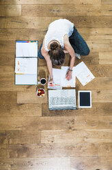 Student sitting on wooden floor surrounded by papers, laptop, digital tablet, file folder, coffee and fruit bowl - UUF004702