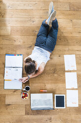Student lying on wooden floor surrounded by papers, laptop, digital tablet, file folder, coffee and fruit bowl - UUF004742