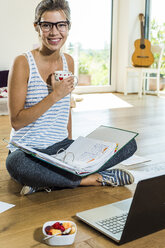 Lächelnde junge Frau auf Holzboden sitzend mit Aktenordner und Laptop - UUF004745