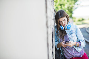 Brünette junge Frau mit Kopfhörern und Handy an die Wand gelehnt - UUF004721