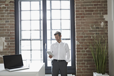 Geschäftsmann am Fenster mit Blick auf ein digitales Tablet, lizenzfreies Stockfoto