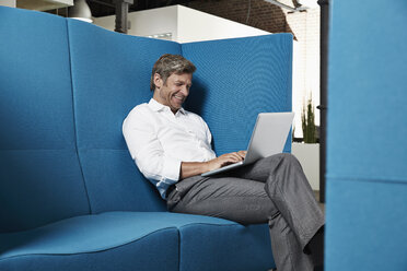 Lächelnder Geschäftsmann, der in einer Gesprächsgrube im Büro sitzt und einen Laptop benutzt - PDF001027