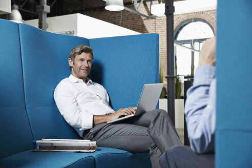 Businessman sitting in conversation pit in office looking at colleague - PDF001028