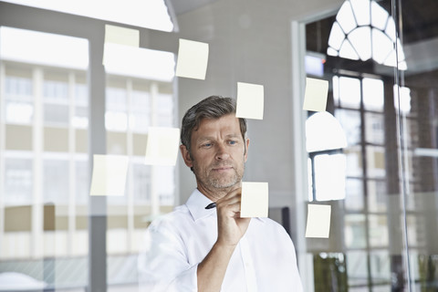 Geschäftsmann schreibt auf Haftnotiz auf Glasscheibe im Büro, lizenzfreies Stockfoto