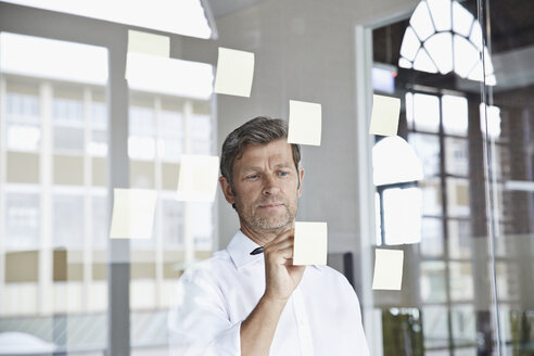 Businessman writing on sticky note on glass pane in office - PDF001046