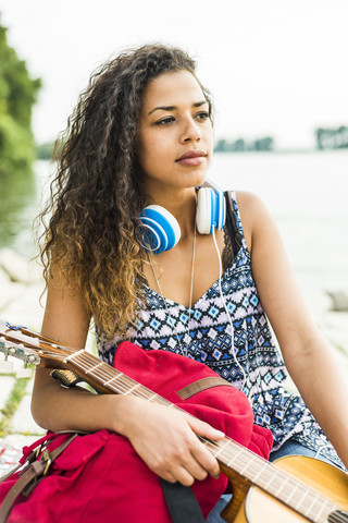 Junge Frau mit Kopfhörern, Gitarre und Rucksack am Flussufer, lizenzfreies Stockfoto