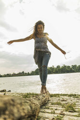 Junge Frau balanciert auf einem Baumstamm am Flussufer - UUF004801