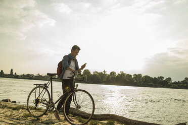 Junger Mann mit Fahrrad und Mobiltelefon am Flussufer - UUF004815