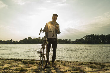 Young man with bicycle and cell phone by the riverside - UUF004816