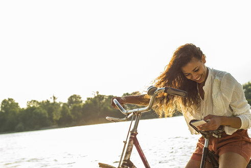 Smiling young woman with bicycle and cell phone by the riverside - UUF004837