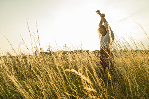 Junge Frau steht mit erhobenen Armen in einem Feld mit hohem Gras - UUF004830