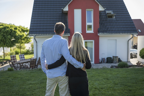 Ein glückliches Paar steht Arm in Arm im Garten und schaut auf sein Haus, lizenzfreies Stockfoto