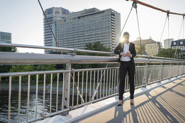 Businesswoman standing on a bridge at twilight using smartphone - PAF001444