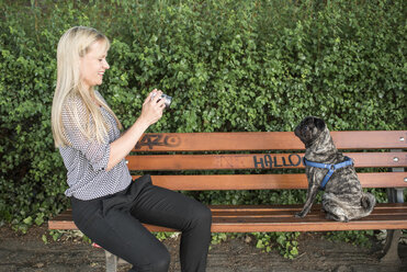 Smiling woman sitting on a bench taking a photo of her pug - PAF001446