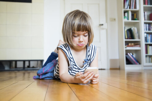 Little girl lying on wooden floor looking sceptical at smartphone - LVF003532