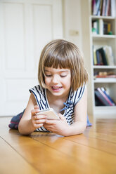 Portrait of smiling little girl lying on wooden floor with smartphone - LVF003526