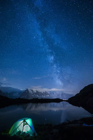 Frankreich, Mont Blanc, Cheserys-See, beleuchtetes Zelt am Seeufer bei Nacht mit Milchstraße und Mont Blanc, der sich im See spiegelt, lizenzfreies Stockfoto