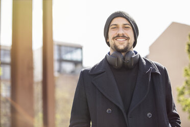 Porträt eines lächelnden jungen Mannes mit Kopfhörern, der eine Wollmütze und einen schwarzen Pullover trägt - TAM000216