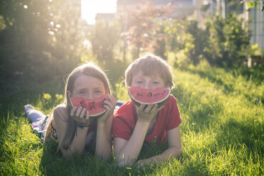Junge und Mädchen liegen auf einer Wiese und bedecken einen Teil ihres Gesichts mit einer Scheibe Wassermelone - SARF001994