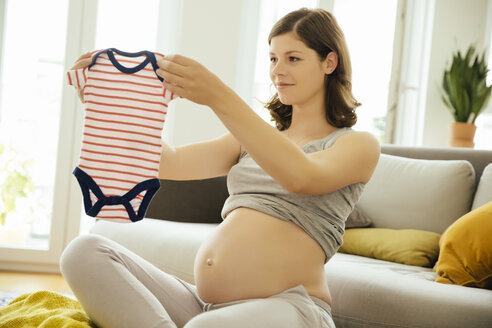 Schwangere Frau hält Babykleidung zu Hause hoch - MFF001666