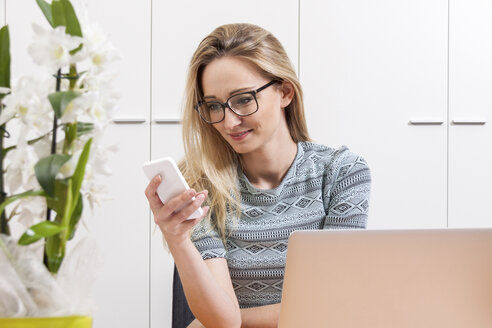 Lächelnde blonde Frau schaut in einem Büro auf ihr Smartphone - JUNF000365