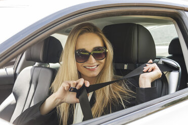 Porträt einer lächelnden blonden Frau mit Sonnenbrille, die in ihrem Auto den Sicherheitsgurt anlegt - JUNF000375