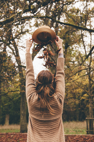 Rückenansicht einer jungen Frau, die sich im Park getrocknete Blätter aus einem Hut auf den Kopf fallen lässt, lizenzfreies Stockfoto
