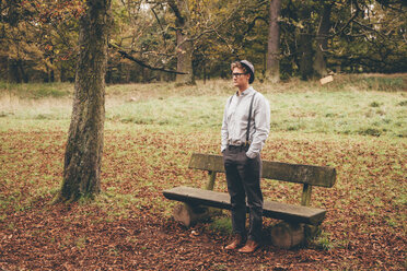 Young man standing in front of a bench in autumnal park looking at distance - CHAF000222