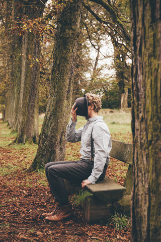 Junger Mann sitzt auf einer Bank in einem herbstlichen Park und bedeckt sein Gesicht mit einem Hut, lizenzfreies Stockfoto