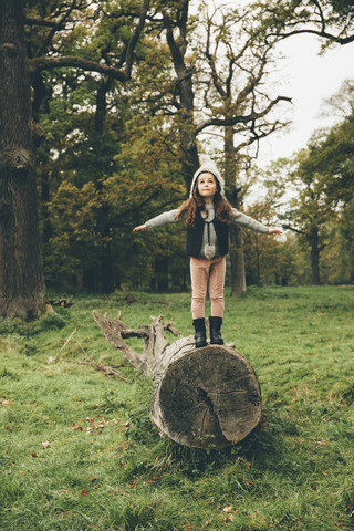 Kleines Mädchen mit Mütze steht mit ausgestreckten Armen auf einem Baumstamm in einem herbstlichen Park, lizenzfreies Stockfoto