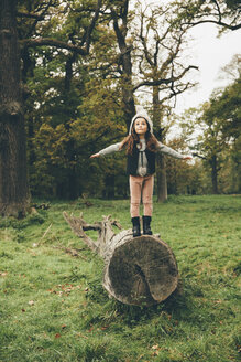 Kleines Mädchen mit Mütze steht mit ausgestreckten Armen auf einem Baumstamm in einem herbstlichen Park - CHAF000237